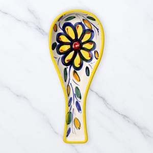 May include: A ceramic spoon rest with a yellow border and a floral design. The flower is yellow with a red center and blue accents. Green and orange leaves and stems decorate the white background.