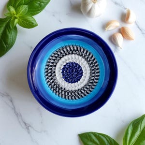 Peut inclure: Un presse-ail en céramique bleu avec des cercles concentriques de différentes nuances de bleu, blanc et noir. Le centre présente un motif texturé bleu foncé. Des feuilles de basilic frais et des gousses d'ail sont disposées autour.