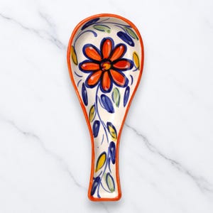 Puede incluir: Un reposacucharas de cerámica con un diseño floral. El reposacucharas tiene una base blanca con una gran flor naranja en el centro, rodeada de hojas azules y detalles amarillos. El borde está delineado en naranja.