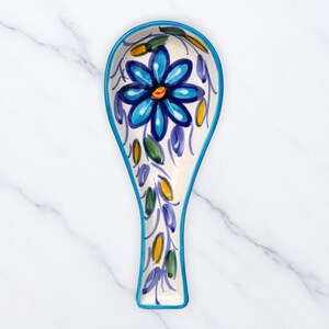 Puede incluir: Un reposacucharas de cerámica con base blanca y diseño floral azul. El reposacucharas tiene un borde azul y está decorado con una gran flor azul, hojas verdes y detalles amarillos. El reposacucharas mide aproximadamente 23 cm de largo.