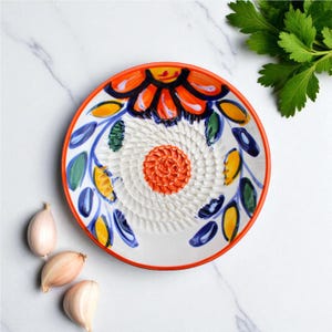 Puede incluir: Un rallador de ajo de cerámica blanca con un diseño floral vibrante. El plato tiene un borde naranja y una superficie de rallado texturizada. Hojas y pétalos pintados en azul, amarillo y verde decoran el borde. Tres dientes de ajo se colocan cerca del plato.