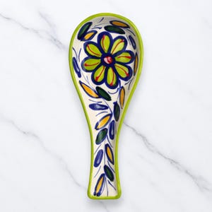 Puede incluir: Un reposacucharas de cerámica con un diseño floral. El reposacucharas tiene una base blanca con un borde verde. El diseño floral presenta una gran flor verde y azul con detalles rojos, rodeada de hojas azules, amarillas y verdes.