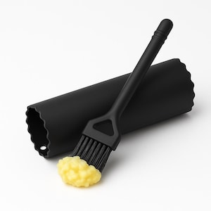 Op de afbeelding: Een zwarte siliconen bakkwast met een gele substantie op de borstelharen, naast een zwarte cilindrische houder. De kwast heeft een lange steel en een driehoekige kop. De houder heeft een geschulpte rand.