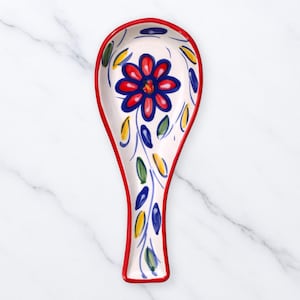 Puede incluir: Un reposacucharas de cerámica con borde rojo e interior blanco. El reposacucharas está decorado con una gran flor roja y azul en el centro, rodeada de diseños de hojas azules, amarillas y verdes.
