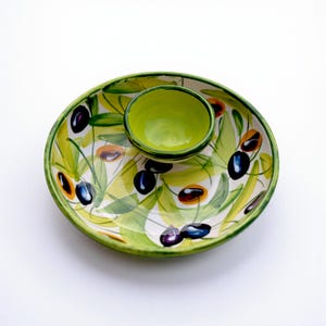 Può includere: Piatto da portata ovale in ceramica con una piccola ciotola rotonda al centro. Il piatto e la ciotola sono dipinti con foglie verdi, olive nere e marroni e un bordo verde. Lo sfondo è bianco.