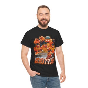 Op de afbeelding: Zwart T-shirt met een afbeelding van de mascotte Gritty en andere personages. De afbeelding is in oranje, rode en bruine tinten. Het woord "GRITTY" is in oranje en wit gedrukt. Het T-shirt wordt door een persoon gedragen.