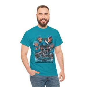 Könnte beinhalten: Türkisfarbenes T-Shirt mit einem Hockey-Team-Grafik und dem Wort "Celebration". Die Grafik zeigt Hockeyspieler in Blau und Weiß, mit einer Trophäe in der Mitte. Ein lässiges Kleidungsstück.