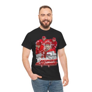 Könnte beinhalten: Schwarzes T-Shirt mit einer Grafik von Eishockeyspielern in roten Trikots. Das Design enthält den Text "Detroit Red Wings" in einer stilisierten roten Schriftart mit einem Stadionhintergrund. Das Shirt ist aus weichem Material.