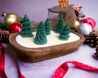 May include: A wooden bowl candle with a white wax base and five green pine tree candle decorations. The bowl is on a white surface with a red ribbon.