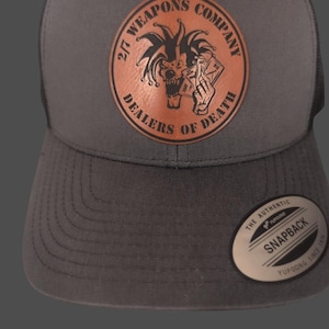 May include: A gray and black snapback hat with a leather patch featuring a jester skull graphic and the text "2/7 WEAPONS COMPANY DEALERS OF DEATH". The hat has a curved brim and a mesh back.