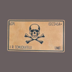 May include: A rectangular, tan-colored patch with a skull and crossbones emblem. The patch has the text "LCPL", "IS12345A+", "I. R. SCHUCKATELLI", and "UNIT" printed in dark blue.