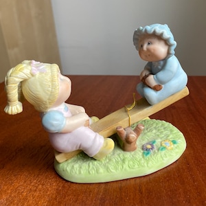 May include: A porcelain figurine of two children on a seesaw. The girl has blonde hair and is wearing a pink dress. The boy has blue hair and is wearing a blue shirt. The seesaw is made of wood and has a green base.