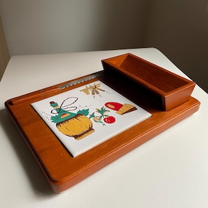 May include: A wooden cheese board with a ceramic tile insert featuring a wine bottle, grapes, and cheese. The board has a small side compartment for utensils.