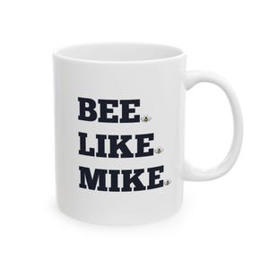 May include: White ceramic mug with the text "BEE LIKE MIKE" in bold, black letters. Small cartoon bees are placed next to each word. The mug has a curved handle.
