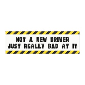 May include: A rectangular sign with the text "NOT A NEW DRIVER JUST REALLY BAD AT IT" in bold black letters. The sign has a white background with black and yellow diagonal stripes along the top and bottom edges.