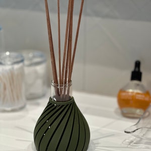 May include: A dark green diffuser bottle with a unique swirl design and wooden sticks. The bottle is sitting on a white surface.