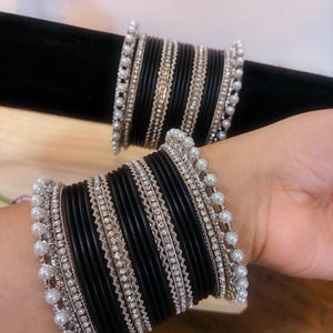 May include: Black bangles with silver and pearl detailing. The bangles are displayed on a wrist and a black velvet surface. The silver accents feature small rhinestones and a pearl border.