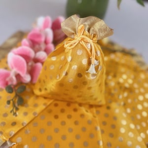 Può includere: Un piccolo sacchetto giallo con coulisse, pois dorati e una parte superiore dorata. La borsa è legata con un nastro giallo e ha piccole nappe argentate. La borsa è su un tessuto giallo con pois dorati.