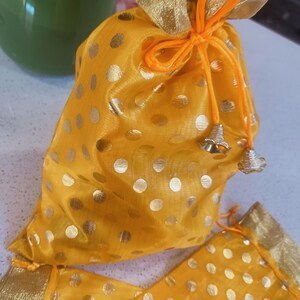 Puede incluir: Una bolsa de regalo amarilla con lunares dorados y un ribete dorado. La bolsa está atada con un cordón naranja y tiene pequeños dijes de campana dorados. La bolsa está hecha de una tela transparente.