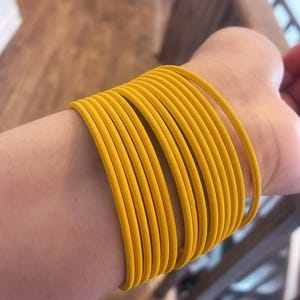以下が含まれることがあります： 黄色いバングルが手首に重ね付けされたクローズアップ。バングルは鮮やかな単色の黄色で、しなやかな素材でできているようです。ブレスレットは互いに密接に積み重ねられており、大胆で人目を引く外観を作り出しています。