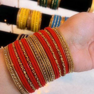 Peut inclure: Une collection de bracelets colorés. L'avant-plan présente une pile de bracelets rouges avec des ornements dorés. D'autres bracelets jaunes, verts et bleus sont visibles à l'arrière-plan, exposés sur du velours noir.