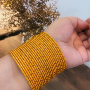 May include: A collection of yellow bangles with gold detailing encircles a wrist. The bangles are a bright mustard yellow and seem to be made of metal or a similar material. The gold flecks provide a subtle shimmer to the jewellery.