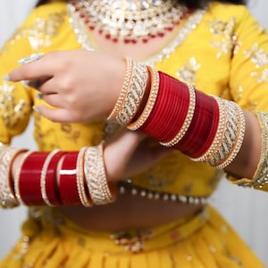 Puede incluir: Un conjunto de pulseras rojas y doradas. Las pulseras están apiladas en la muñeca, con bandas alternas rojas y doradas. Las bandas doradas están decoradas con pequeñas piedras brillantes. El fondo es una prenda amarilla con bordados dorados.