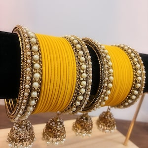 May include: A collection of yellow bangles with pearl and gold detailing. The bangles are stacked, with a decorative gold charm suspended from each. The bangles are presented on a wooden surface.