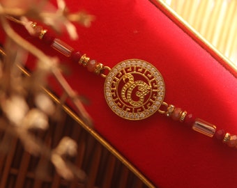 May include: Gold-toned bracelet with a circular pendant featuring a stylized design, encrusted with small, clear stones. The bracelet has red and gold beads and is set against a red background.