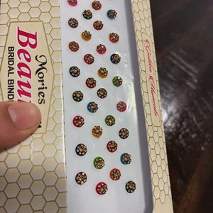 May include: A box of Mories Bridal Bindis, featuring a variety of colorful, jewel-toned forehead adornments. Each bindi has a central cluster of small, sparkling gems surrounded by a colored base. The box has a geometric pattern.