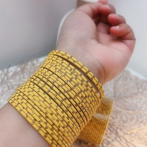 Op de afbeelding: Een stapel gele armbanden met gouden glitteraccenten siert een pols. De armbanden hebben een levendige gele kleur en zijn dicht op elkaar gestapeld. De afbeelding toont ook een vaas met roze bloemen.