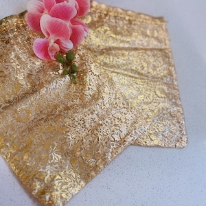 May include: A gold-colored, patterned fabric with a floral design is displayed. A cluster of pink orchids with green stems rests on the fabric. The fabric appears to be folded, creating a textured surface.