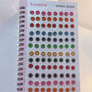 May include: A spiral-bound book of Ananya Kumkum Bindi, featuring rows of colorful, circular bindis. The bindis are in various colors, including red, orange, blue, green, and pink, each with a gold-colored border and a central jewel.