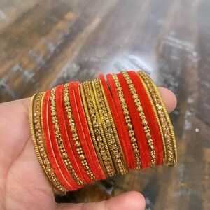 May include: A collection of red and gold bangles. The bangles are a vivid red with gold detailing and a textured pattern. The gold elements feature small, decorative beads and intricate designs. The bangles are stacked, highlighting their design.