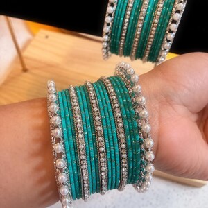 May include: Teal bangles with silver detailing and faux pearls. The bangles are stacked, creating a layered effect. The silver detailing includes a row of small, clear stones. The pearls are white and round.