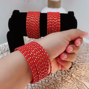 Peut inclure: Ensemble de bracelets rouges ornés de points dorés. Les bracelets sont présentés sur un présentoir noir et portés au poignet. L'image comprend un vase avec des fleurs roses.