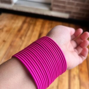 Peut inclure: Une pile de bracelets roses vifs entoure un poignet. Les bracelets sont d'un rose fuchsia vibrant et semblent être faits d'un matériau lisse et solide. L'arrière-plan est une vue floue d'une surface en bois et d'un mur de briques.