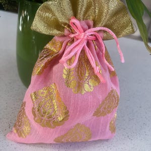 Può includere: Un sacchetto regalo in tessuto rosa con motivi floreali dorati e chiusura con coulisse dorata. La parte superiore della borsa è arricciata con un tessuto dorato e scintillante. La borsa è legata con uno spago rosa.