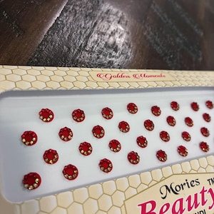 Op de afbeelding: Een verpakking rode en gouden bindi's, klein, rond en juweelachtig, gerangschikt in rijen. De verpakking is wit met een honingraatpatroon en de woorden "Golden Moments" en "Mories Beauty Bridal Bindi".