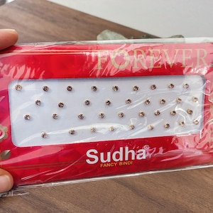 May include: A red packet of Sudha Fancy Bindi, containing numerous small, round, brown decorative dots. The package features the word "FOREVER" in gold lettering and a floral design.
