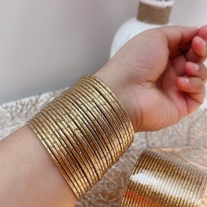 May include: A stack of gold-colored bangles adorns a wrist, with a matching set resting nearby. The bangles have a textured surface and a metallic sheen. The background includes a decorative surface and a vase with pink flowers.