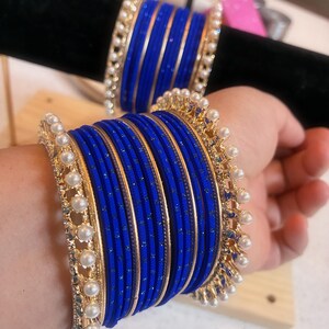 Puede incluir: Un conjunto de brazaletes azules vibrantes con detalles dorados y adornos de perlas de imitación. Los brazaletes están apilados, mostrando su intrincado diseño y color. Las joyas se exhiben sobre una superficie negra.