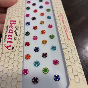 May include: A package of Mories Beauty Bridal Bindi, featuring a variety of colorful, jewel-like forehead adornments. The bindis are in various colors, including red, pink, green, blue, and orange, with a central gold-colored accent.