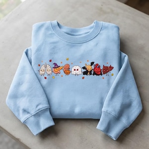 Peut inclure: Un sweat-shirt bleu clair à col rond orné d'un motif central représentant des organes humains en dessin animé déguisés pour Halloween, incluant une momie, une sorcière, une citrouille, un fantôme, un vampire et un diable, avec des feuilles d'automne.