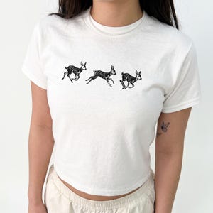 Può includere: Una t-shirt bianca a maniche corte con una stampa grafica nera di tre piccoli cerbiatti in movimento sul petto. La maglietta presenta un girocollo e un design semplice e pulito, che mette in risalto le dettagliate illustrazioni dei giovani cervi.