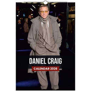 Puede incluir: Calendario de 2026 con Daniel Craig. La imagen muestra a Craig con un traje, abrigo y bufanda de color marrón claro. El calendario tiene una pancarta negra con el nombre "DANIEL CRAIG" y "CALENDAR 2026" en blanco y rojo. El calendario está colgado.