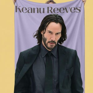 May include: A lavender blanket featuring a portrait of Keanu Reeves with the text "Keanu Reeves" in a dark font. The image shows a man in a black suit, black shirt, and tie.