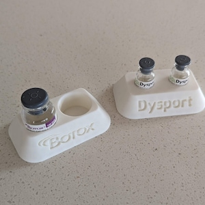May include: Two white plastic stands with the words "Botox" and "Dysport" printed on them. Each stand holds small glass vials with black caps.