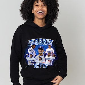 May include: Black hoodie featuring a graphic print of baseball players in blue and white uniforms with the text "Mookie Betts" in blue and white. The background is a blue and purple gradient with stars.