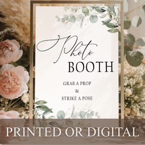 May include: A rectangular sign in a thin gold frame featuring the text Photo BOOTH, GRAB A PROP & STRIKE A POSE in black lettering. The white background is decorated with green eucalyptus leaves and branches at the top and bottom corners.